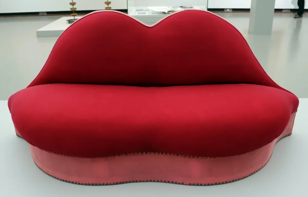 Red lips-shaped sofa by Salvador Dalí, surrealist artwork displayed in a museum.