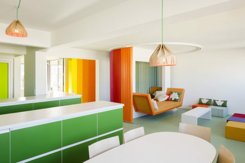 le rideau incurvé composé de lames verticales multicolores. Il s’agit de la structure mobile arc-en-ciel qui permet de moduler l’espace central. Cette vue est emblématique du dispositif “Rainbow”, dont l’intention est de "créer un paysage d’intérieur vivant, dynamique, coloré".