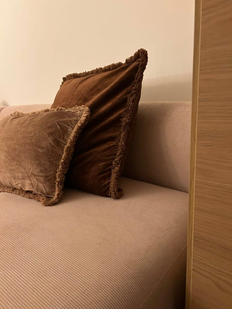 Sklum sofa in powder pink fabric with brown fringed velvet cushions, contemporary interior with clean lines and soft, warm palette.