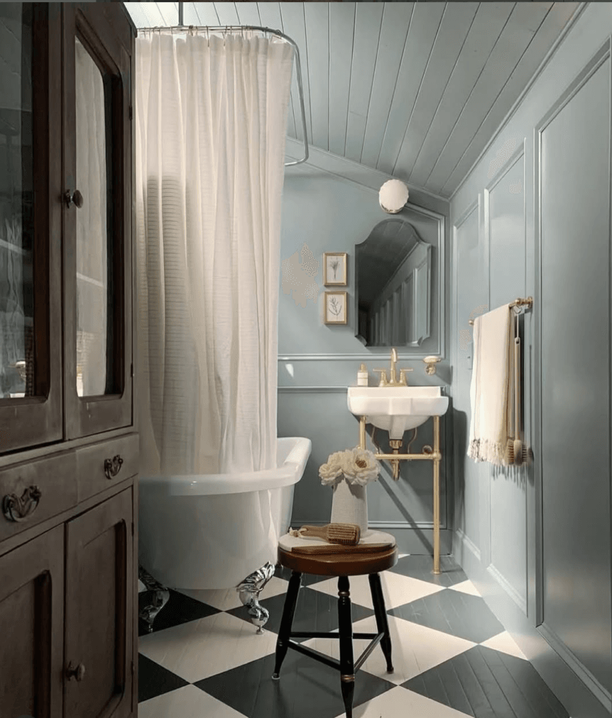 Petite salle de bain d’inspiration cottagecore avec baignoire sur pieds, panneaux muraux bleu pâle, sol à carreaux graphiques noir et blanc, robinetterie en laiton et meuble ancien servant de rangement.

