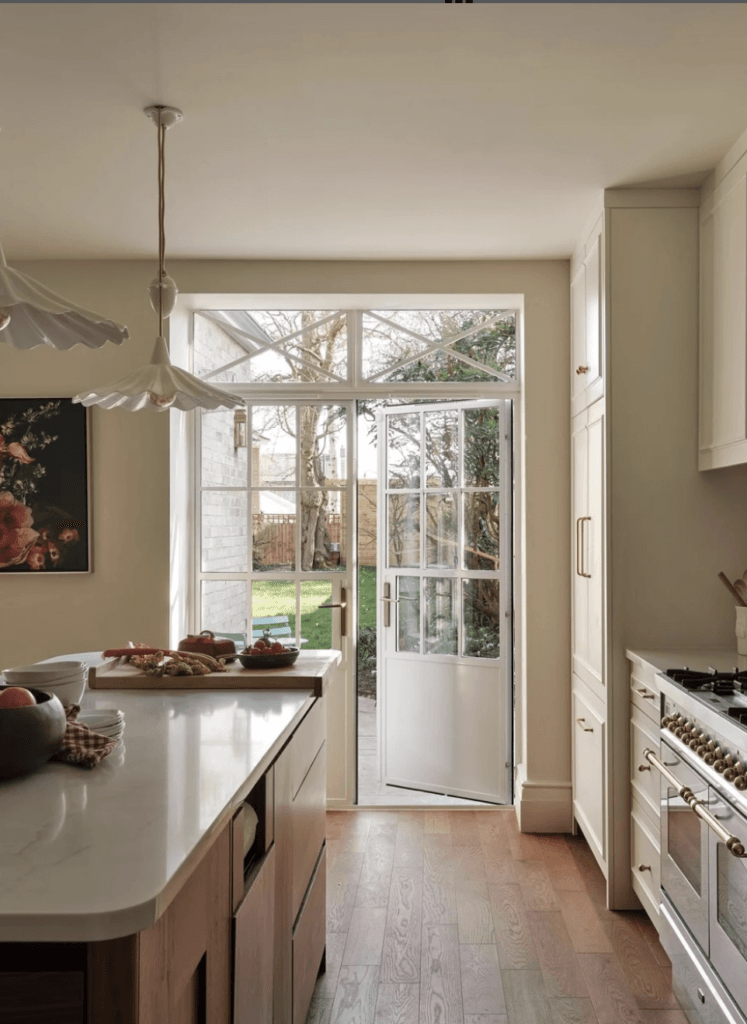 Cuisine lumineuse d’inspiration Arts & Crafts et cottagecore, avec îlot central en noyer aux angles arrondis, lambris clairs, parquet en bois, grandes fenêtres à ossature métallique laissant entrer une lumière traversante, et ouverture sur le jardin.