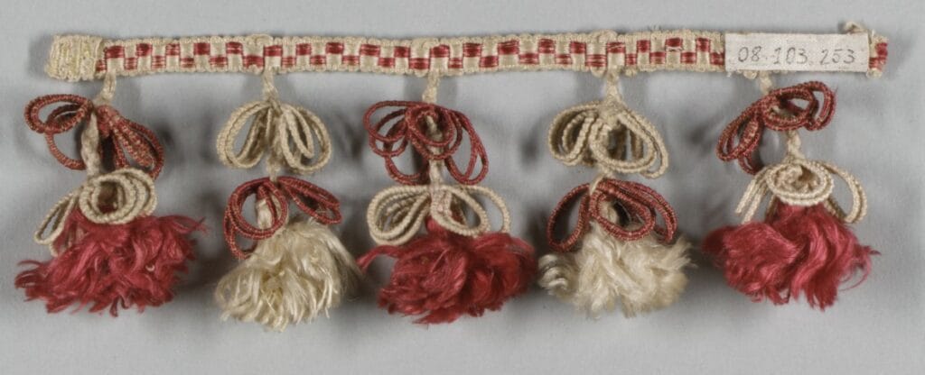 Passementerie d’ameublement du XIXᵉ siècle avec franges et glands en fils rouges et écrus, élément décoratif caractéristique du style Louis-Philippe.