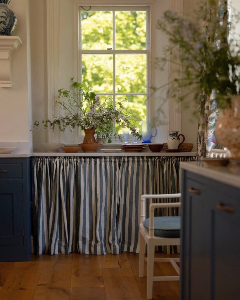 Cuisine inspirée du cottagecore avec meubles bleu grisé, crédence à motifs traditionnels, vaisselle artisanale posée sur le plan de travail et rideaux en tissu fleuri encadrant une fenêtre ouverte sur un jardin verdoyant.